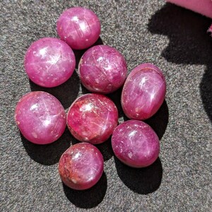 Könnte beinhalten: Eine Sammlung von acht polierten, runden Edelsteinen in verschiedenen Rosa- und Rottönen. Die Steine haben eine glatte, reflektierende Oberfläche mit sichtbaren inneren Einschlüssen. Die Edelsteine sind auf einer dunklen, strukturierten Oberfläche angeordnet.