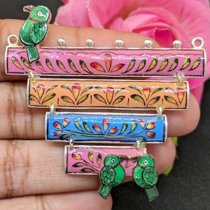 May include: A silver pendant with five colorful rectangular bars, each with a different floral design. The top bar has a green bird perched on it. The bottom bar has two green birds facing each other.