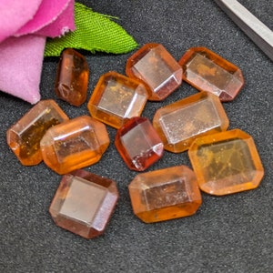 Può includere: Una collezione di dieci gemme di vetro sfaccettate di colore ambrato. Le gemme hanno tutte la stessa forma, ma variano leggermente di dimensioni. Sono disposte in uno schema casuale su una superficie grigio scuro.