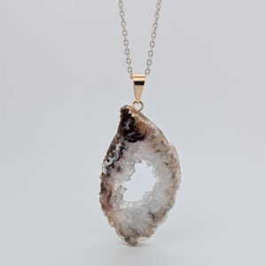 May include: A rose gold chain necklace with a geode slice pendant. The pendant has a natural shape, with a white, crystal-filled centre and brown and grey outer edges. The necklace is shown against a white background.
