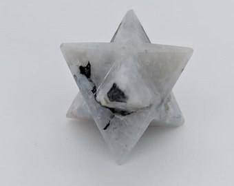 Puede incluir: Una estrella Merkaba de cristal blanco y gris. La estrella de seis puntas tiene inclusiones negras y está facetada. El cristal está sobre un fondo blanco.