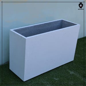 Può includere: Una fioriera rettangolare in cemento bianco con un lato inclinato. L'interno è di colore grigio scuro. La fioriera è progettata per uso esterno ed è posizionata contro un muro azzurro e un prato verde.