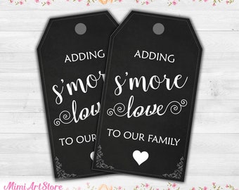 May include: Two black rectangular gift tags with rounded top corners and a hole punch. The tags feature white text that reads "ADDING s'more love TO OUR FAMILY" with a small white heart icon at the bottom and decorative corner flourishes.