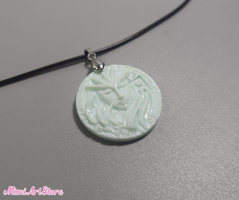 May include: A light blue pendant necklace featuring a carved face design. The round pendant is attached to a black cord with a silver-colored clasp. The Mimi Art Store logo is visible in the bottom left corner.