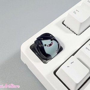 May include: A black and blue keycap with a cartoon character with black hair and a pink mouth. The keycap is on a white keyboard.