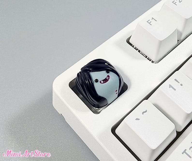 May include: A black and blue keycap with a cartoon character with black hair and a pink mouth. The keycap is on a white keyboard.