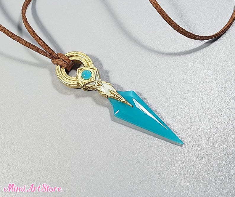 May include: A pendant necklace featuring a turquoise dagger-shaped charm with gold and white accents. The charm is suspended from a brown cord. The necklace has a gold ring clasp. The Mimi Art Store logo is visible in the bottom left corner.