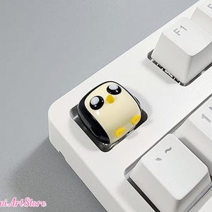 May include: A white keyboard with a black and white penguin keycap. The penguin has black eyes and a yellow beak.