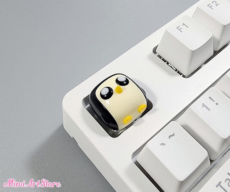 May include: A white keyboard with a black and white penguin keycap. The penguin has black eyes and a yellow beak.