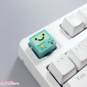 May include: A teal green keycap with a smiling face and a star, triangle, and circle design. The keycap is on a white keyboard.