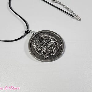 May include: A silver-colored pendant necklace featuring a detailed phoenix design within a circular frame. The pendant hangs from a black cord with a silver-toned chain extender. The necklace is displayed on a white surface.