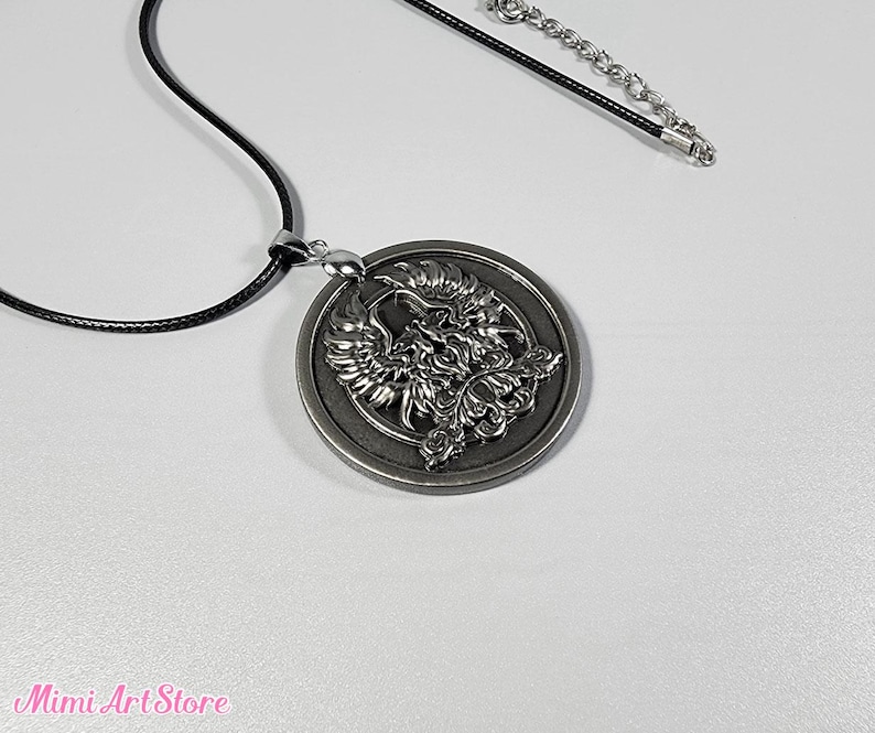 May include: A silver-colored pendant necklace featuring a detailed phoenix design within a circular frame. The pendant hangs from a black cord with a silver-toned chain extender. The necklace is displayed on a white surface.