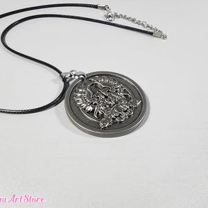 Könnte beinhalten: Halskette mit silberfarbenem Anhänger, der ein detailliertes Phönix-Design zeigt. Der runde Anhänger ist an einer schwarzen Kordelkette mit silberfarbener Verlängerungskette befestigt. Die Halskette liegt auf einer weißen Oberfläche.