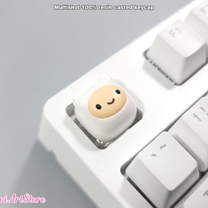 May include: White keyboard with a custom keycap featuring a cartoon character with a white hat and a yellow face. The character has black eyes and a small smile. The keycap is on the "~" key.