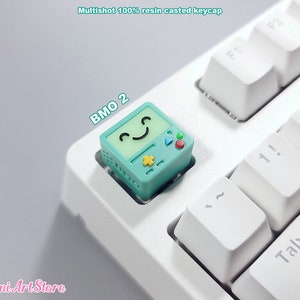 May include: A teal blue resin keycap with a cartoon face and the text "BMO 2" on it. The keycap is on a white keyboard.
