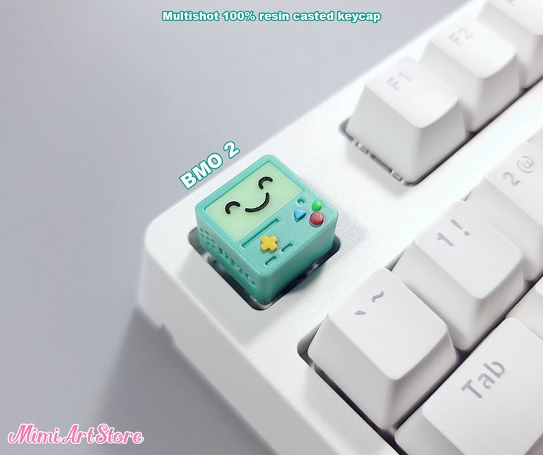 May include: A teal blue resin keycap with a cartoon face and the text "BMO 2" on it. The keycap is on a white keyboard.