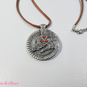 May include: A silver-colored pendant necklace with a detailed dragon design and three red accents. The pendant hangs from a brown cord necklace with a silver clasp. The Mimi Art Store logo is visible in the bottom left corner.