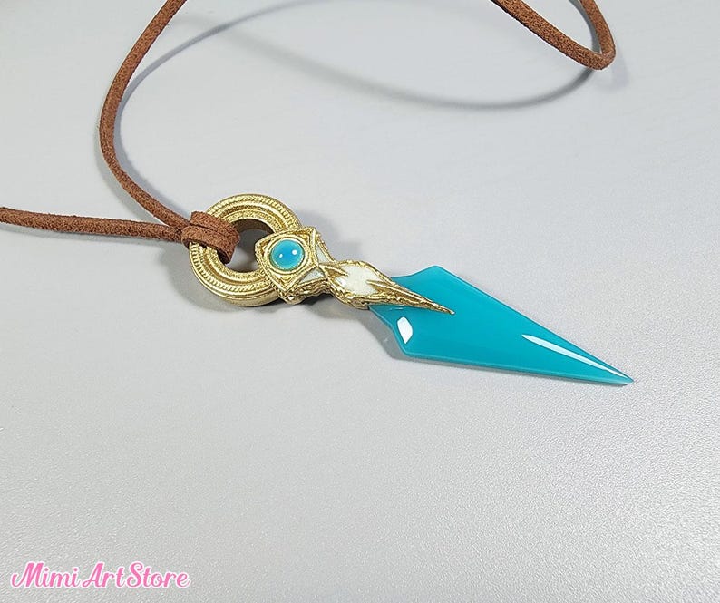 May include: A pendant necklace featuring a turquoise dagger-shaped charm with gold and white accents. The charm is attached to a brown leather cord. The pendant has a gold ring with a turquoise stone.