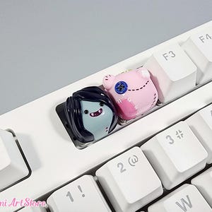 May include: Two small, colorful, 3D printed keycaps featuring characters from the animated series Adventure Time. The keycaps are placed on a white keyboard. The keycaps are made to look like Finn the Human and Marceline the Vampire Queen.