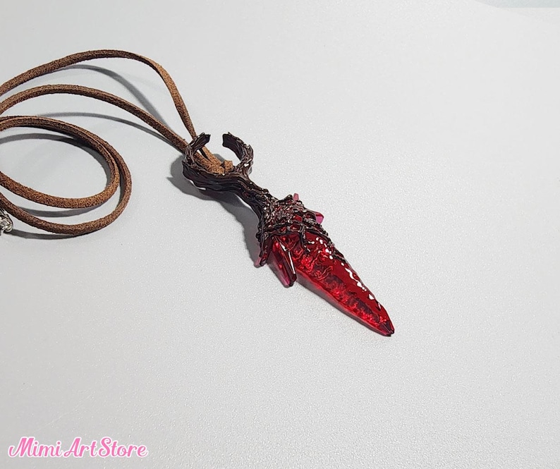 May include: A handcrafted pendant necklace featuring a deep red, dagger-shaped crystal. The crystal is set in a dark brown, textured metal setting and hangs from a brown leather cord. The Mimi Art Store logo is visible in the bottom left corner.