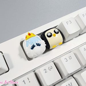 May include: Two small, cartoon character keycaps for a keyboard. The keycaps are made of white, black, and yellow plastic. The first keycap is a penguin with a black body and a white head. The second keycap is a white character with a crown and a sad expression.