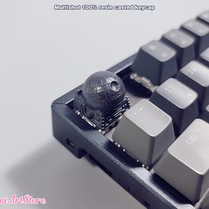 Puede incluir: Un tapón de tecla de resina negro y gris con un diseño detallado que se asemeja a la Estrella de la Muerte de Star Wars. El tapón de tecla está en un teclado negro.