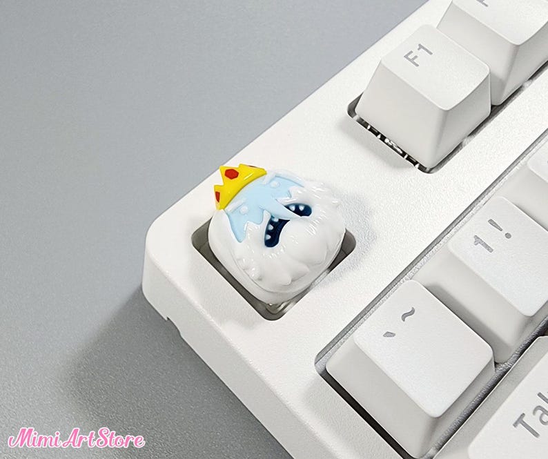 May include: A white and blue ice king character with a yellow crown, designed as a keycap for a keyboard.