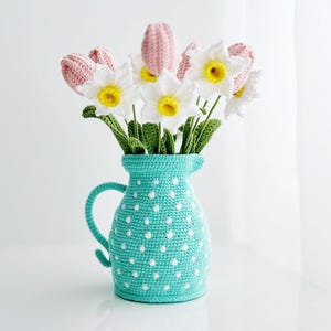 Op de afbeelding: Een gehaakte kan met witte stippen op een turquoise achtergrond bevat een boeket van gehaakte roze tulpen en witte narcissen.
