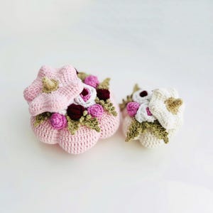 Peut inclure: Deux contenants en forme de fleur au crochet. L'un est rose clair avec un couvercle assorti et est décoré de fleurs roses, bordeaux et blanches. L'autre est blanc avec un centre beige et est également décoré de fleurs. Les contenants sont sur une surface blanche.