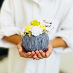 May include: A gray crocheted sphere with a bouquet of yellow, white, and green crocheted flowers on top. The sphere is being held in the hands of a person. A small white card with the text "Thank you!" is visible in the background.