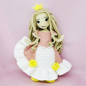 Pode incluir: Boneca de crochê feita à mão em forma de princesa. A boneca tem cabelo loiro comprido, uma coroa amarela e um vestido rosa e branco. Rosas amarelas são presas à saia. A boneca tem aproximadamente 30 cm de altura.