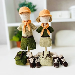 Puede incluir: Dos muñecas de crochet vestidas con uniformes de exploradores con mochilas y botas a juego. Las muñecas llevan gorros naranjas y ropa verde y marrón.