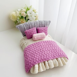 May include: A miniature crochet bed set with a gray headboard, two pink square pillows, and a pink bolster pillow. The bed has a white and pink blanket with a ruffled edge. A bouquet of white and yellow flowers is in the background.