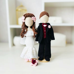 May include: Crocheted bride and groom dolls, the bride is wearing a white dress with a pink flower crown and the groom is wearing a black suit with a red bow tie.