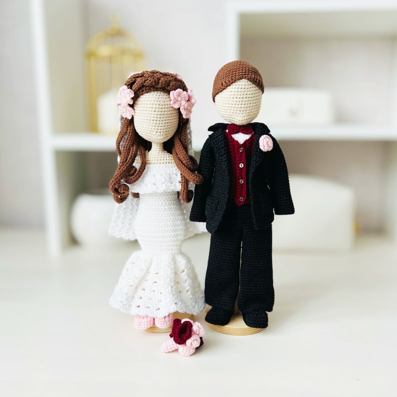 May include: Crocheted bride and groom dolls, the bride is wearing a white dress with a pink flower crown and the groom is wearing a black suit with a red bow tie.