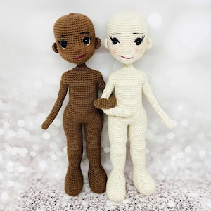 Puede incluir: Dos muñecas de crochet, una con piel marrón y otra con piel blanca, de pie una al lado de la otra. Ambas muñecas tienen ojos negros y labios rojos. La muñeca con piel marrón lleva un atuendo marrón, y la muñeca con piel blanca lleva un atuendo blanco.
