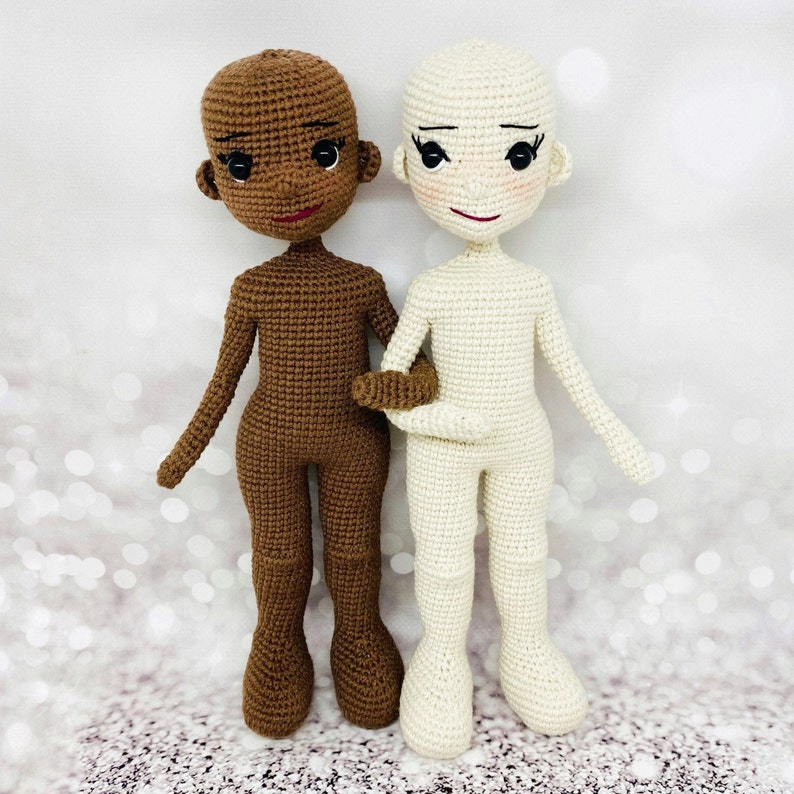 May include: Two crocheted dolls, one with brown skin and one with white skin, standing side by side. Both dolls have black eyes and red lips. The doll with brown skin is wearing a brown outfit, and the doll with white skin is wearing a white outfit.