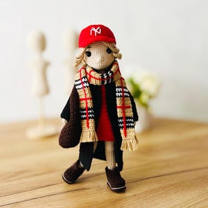 May include: A crocheted doll wearing a red and black plaid scarf, a red hat with the letter "N" on it, and a brown coat. The doll is standing on a wooden surface.