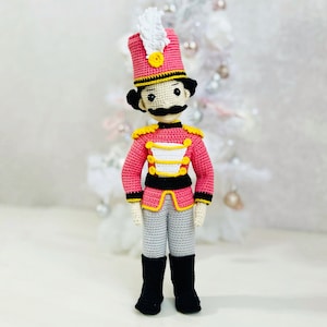 May include: A handmade crochet toy soldier in a pink uniform with gold accents, black boots, and grey trousers. The soldier has a black moustache and a tall pink hat with a white feather. A charming, decorative item.