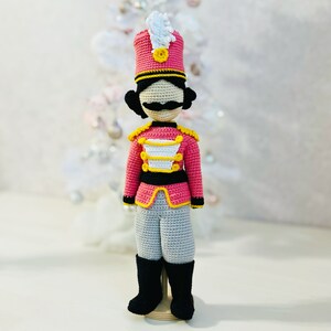 Puede incluir: Un soldado cascanueces de crochet que lleva un uniforme rojo y blanco con botones dorados y un sombrero negro con una pluma blanca. El juguete está de pie sobre una base de madera.