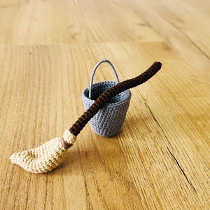 以下が含まれることがあります： 茶色の柄とベージュのほうき頭が付いた、かぎ針編みのミニチュアほうき。小さな灰色の、持ち手が付いたかぎ針編みのバケツの隣にあります。木製の表面に置かれています。