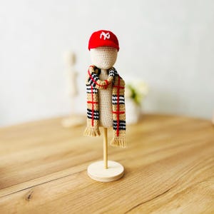 Op de afbeelding: Een gehaakte pop met een rode baseballpet met een wit "NY" logo en een tan en zwart geruite sjaal. De pop staat op een houten standaard.