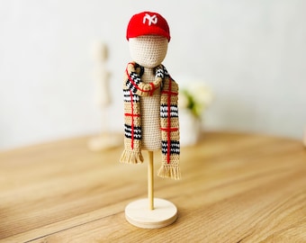 May include: A crocheted doll wearing a red baseball cap with a white "NY" logo and a tan and black plaid scarf. The doll is on a wooden stand.