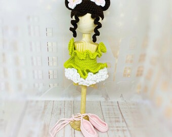May include: A crocheted doll with brown hair, green and white dress, and pink ballet shoes. The doll is standing on a wooden stand.