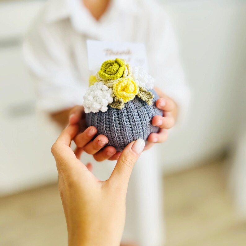 May include: A gray crocheted ball with a bouquet of yellow, white, and green crocheted flowers on top. The ball has a tag that says "Thank You".