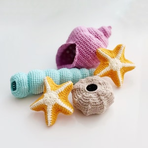 May include: A collection of five crocheted sea life items including a light purple conch shell, a long light blue cylindrical shell, two bright yellow starfish with white centres, and a round textured beige sea urchin with a dark centre hole.