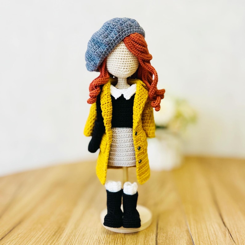 May include: A crocheted doll wearing a yellow coat, a black sweater, a white collared shirt, a gray skirt, black boots, and a blue beret. The doll has red hair and is standing on a wooden base.
