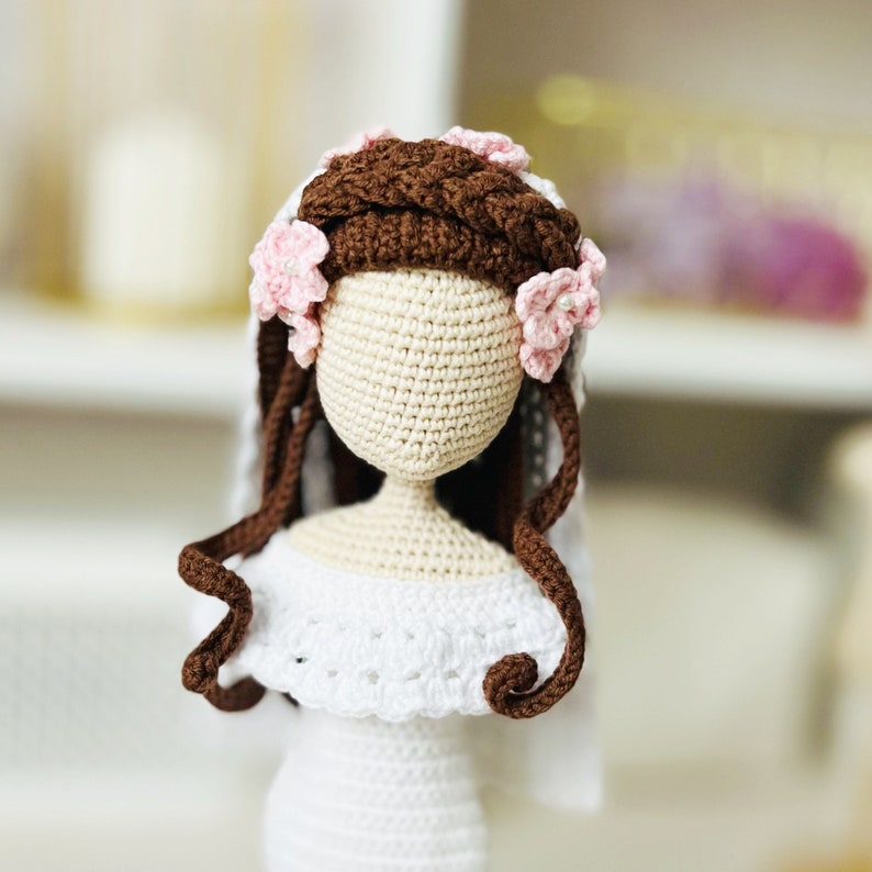 May include: Crocheted doll with brown hair and a white dress. The doll is wearing a pink flower crown.