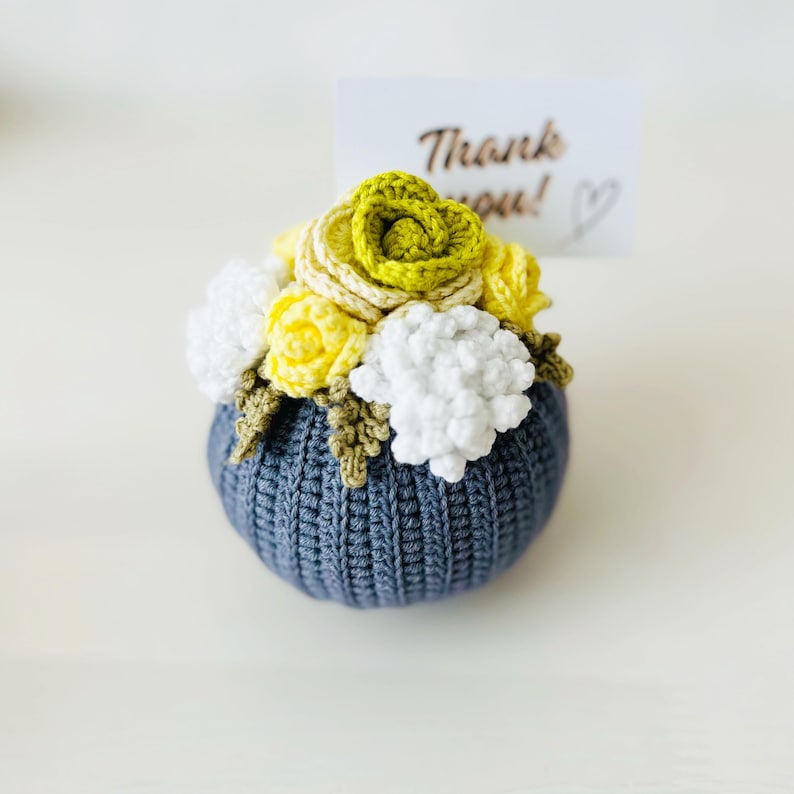 May include: A crocheted flower arrangement in a gray pot. The flowers are yellow, white, and green. The pot has a textured surface. A small card with the text "Thank you!" is behind the arrangement.