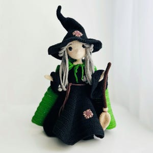 Könnte beinhalten: Gehäkelte Hexenpuppe mit schwarzem Kleid, Hut und grünem Umhang. Die Puppe hat graues Haar, eine rosa Blume auf dem Hut und hält einen kleinen Besen. Eine handgefertigte Halloween-Dekoration.